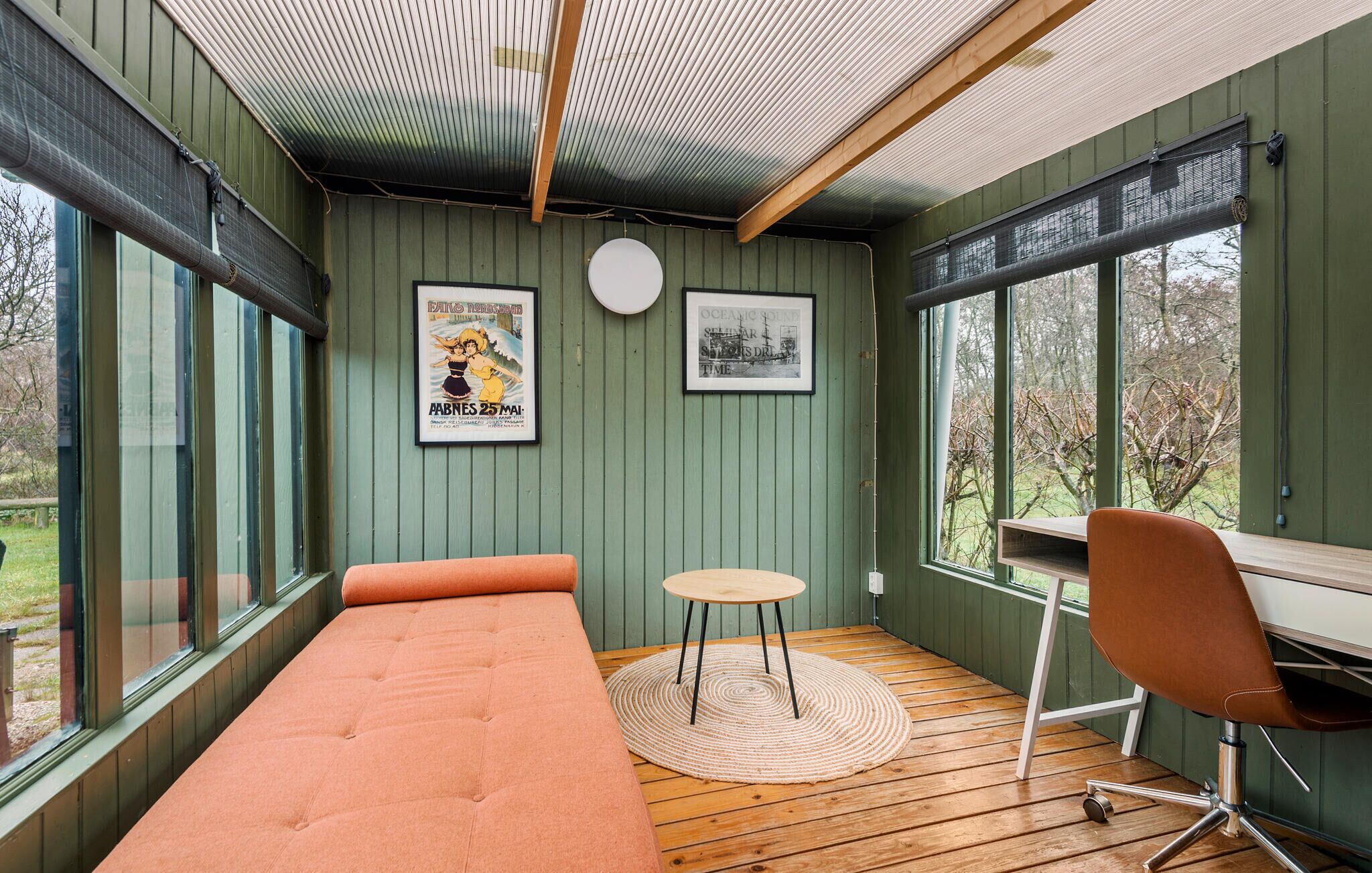A room with a bed, a small round table, a chair, and a desk. There are two large windows and a door.