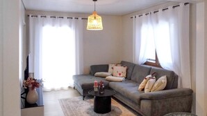 Una sala de estar con un sofá gris, una mesa redonda de centro, una lámpara colgante y un florero con flores.