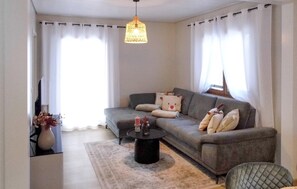 Una sala de estar con un sofá gris, una mesa redonda de centro, una lámpara colgante y un florero con flores.