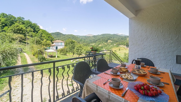 Un balcone con un tavolo apparecchiato per la colazione, affacciato su un paesaggio rurale con una casa e montagne.