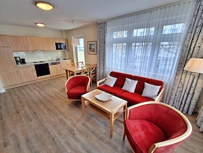 A modern living room with a red sofa, two red armchairs, a wooden coffee table, and a dining area with a table and chairs. A kitchen with wooden cabinets and a microwave is visible in the background. - Holiday Island beach Apartment 3 26 - Apartment 3 26 "island beach" (Heringsdorf)