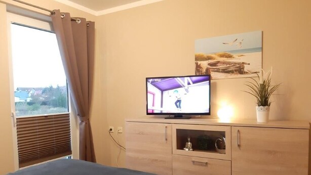A bedroom with a bed, a TV on a wooden cabinet, a window with curtains, and a painting on the wall. - (282) 2-room apartment-Hafenstraße - Apartment complex Yachthafenresidenz (Kühlungsborn)