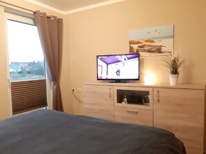 A bedroom with a bed, a TV on a wooden cabinet, a window with curtains, and a painting on the wall. - (282) 2-room apartment-Hafenstraße - Apartment complex Yachthafenresidenz (Kühlungsborn)