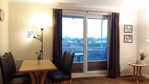 A room with a dining table, chairs, a lamp, a small table with a plant, and a view through a sliding door. - (282) 2-room apartment-Hafenstraße - Apartment complex Yachthafenresidenz (Kühlungsborn)