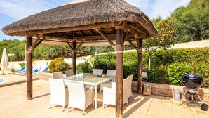 A covered outdoor dining area with a table and chairs, a barbecue grill, and a pool in the background. - Beach front home in Marbella (Marbella)