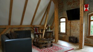 Een loft-achtige kamer met zichtbare houten balken, een zwarte leren bank, een houten salontafel en een rood tapijt met dessin.