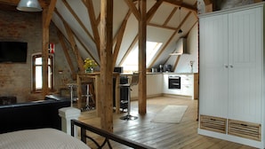 Een slaapkamer in loft-stijl met zichtbare houten balken, een groot bed, een bureau en een kitchenette op de achtergrond.