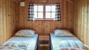 Een hutje met twee eenpersoonsbedden, houten wanden, een raam met ruitjesgordijnen, en een kleine tafel tussen de bedden.