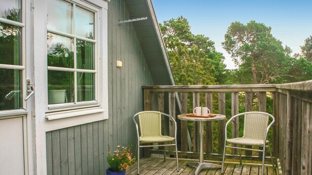 Een houten terras met twee rieten stoelen en een kleine tafel, een plant in pot, en een raam met uitzicht op bomen.