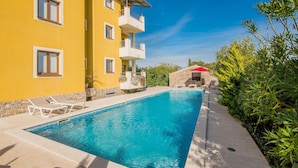 Una piscina con sillas de descanso, un edificio amarillo con balcones y un muro de piedra.
