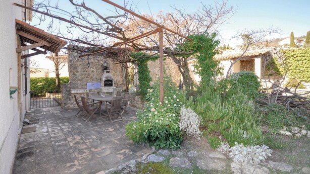 An outdoor patio with a stone floor, a wooden pergola, a stone oven, and surrounding greenery.
