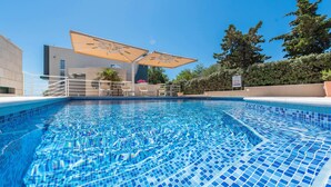 Ein Schwimmbecken mit gefliestem Boden, Sitzgelegenheiten im Freien und Sonnenschirmen.