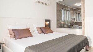Ein modernes Schlafzimmer mit einem großen Bett, zwei Kissen, einem Nachttisch, einer Wand-Klimaanlage und einem großen Spiegel, der ein Badezimmer mit Dusche reflektiert.