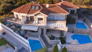 Una casa de varios pisos con fachada de piedra, dos piscinas y un porche cubierto.