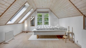 A bedroom with a sloped ceiling, a bed with white bedding, a radiator, and a window with a view of greenery.