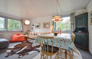 A cozy dining area with a table set for four, surrounded by wooden chairs. There is a large window with curtains, a hanging light fixture, and a green cabinet with a decorative item on top.