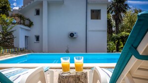 Blick auf einen Pool mit zwei Orangensaftgläsern auf einem Holzstumpf, weißen Liegestühlen und einem Gebäude im Hintergrund, das Klimaanlagen hat.