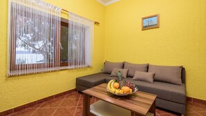 A room with yellow walls, a brown sofa, a wooden table with a fruit bowl, and a window with white curtains. - 1 bedroom beach front apartment in Lukovo Sugarje (Lukovo Sugarje)