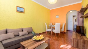 A living room with a sofa, a coffee table with fruit, a dining area with chairs, and a TV stand. - 1 bedroom beach front apartment in Lukovo Sugarje (Lukovo Sugarje)