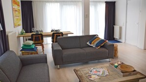 Een moderne woonkamer met een grijze bank, een salontafel met tijdschriften en kaarsen, en een eethoek met een houten tafel en stoelen.