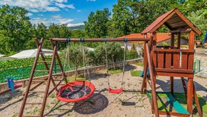 Ein hölzerner Spielplatz mit Schaukeln, einer Taenschaukel und einem kleinen Spielhäuschen.