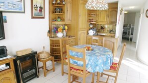 Un coin repas avec une table ronde recouverte d’une nappe bleue, entourée de chaises en bois. On aperçoit un buffet en bois avec des étagères au-dessus de la table, et un panier en osier posé sur le sol.