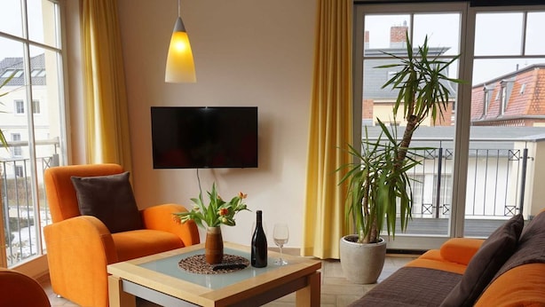 Una sala de estar moderna con muebles naranja, un televisor de pantalla plana, una planta en maceta y vistas de edificios a través de ventanas amplias.