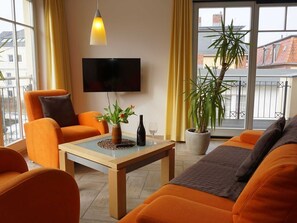 A modern living room with orange furniture, a flat-screen TV, a potted plant, and a view of buildings through large windows. - Apartment 6 - Villa "dunes vis a vis" near the beach (Heringsdorf)