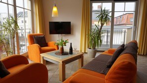 Una sala de estar moderna con muebles naranjas, un televisor de pantalla plana y ventanas grandes con cortinas amarillas.