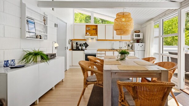 Une cuisine moderne avec des meubles blancs, un coin repas doté de chaises en osier et de grandes fenêtres offrant une vue sur la verdure.