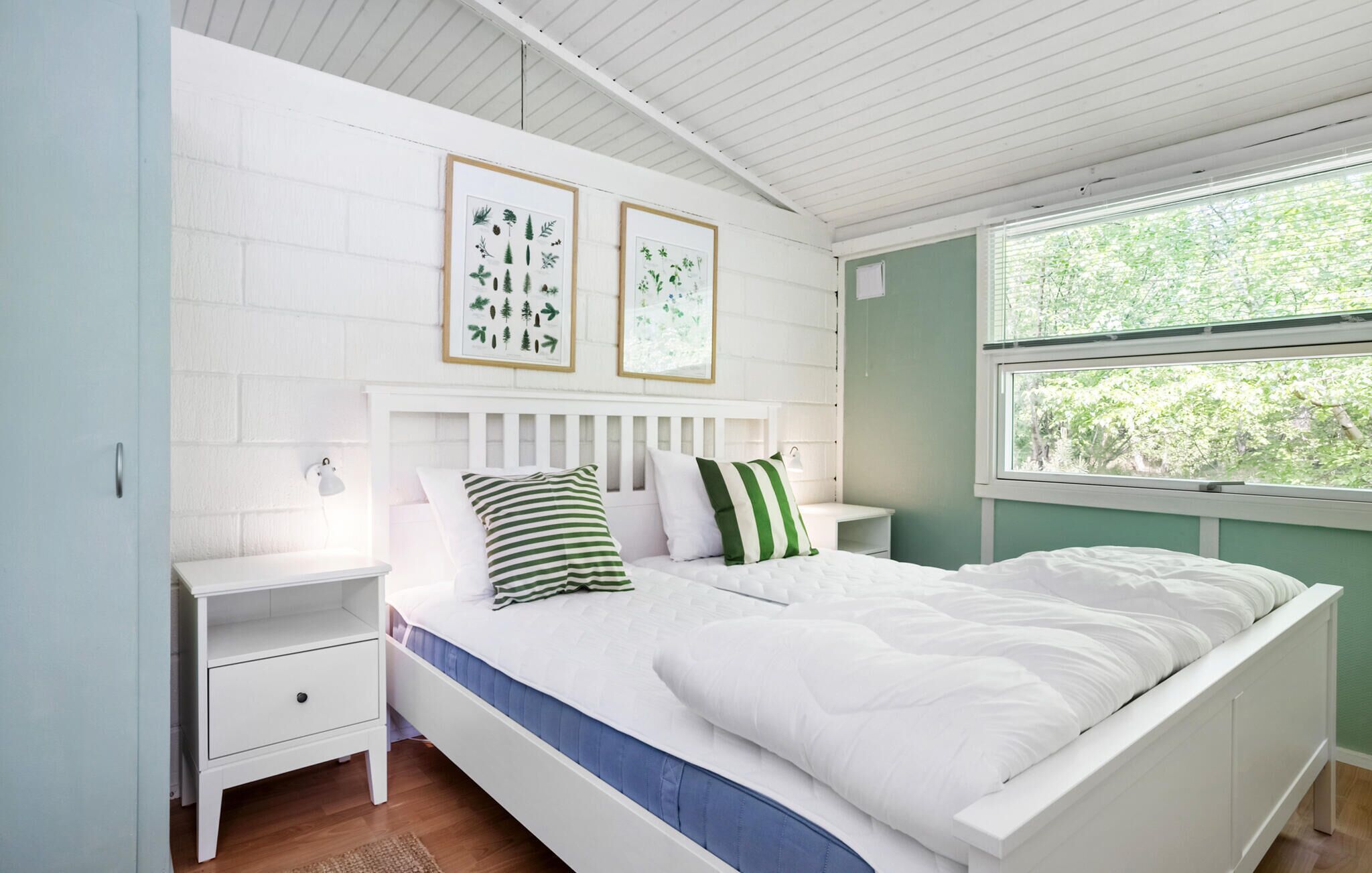 A bedroom with a white bed, green and white striped pillows, a white nightstand, a framed botanical print on the wall, and a window with a view of trees.