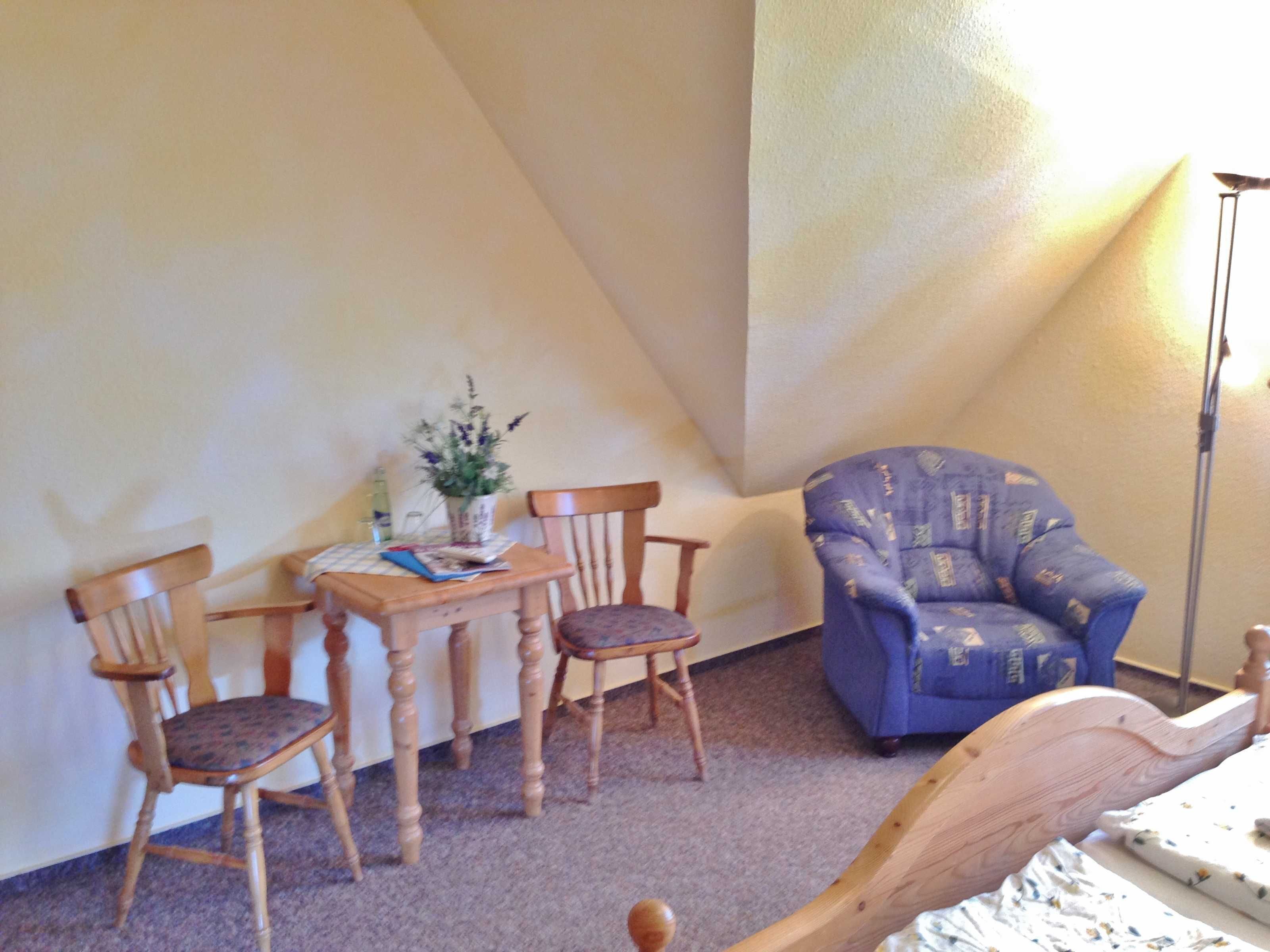 A room with a bed, two wooden chairs, a small table with a vase of flowers, and a blue armchair.
