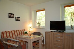 A living room with a striped sofa, a wooden table with a potted plant, a television on a wooden cabinet, and a window with a plant.