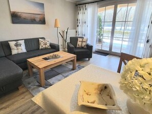 Sala de estar com sofá, mesa de centro e poltrona. Janela com cortinas e vista para o exterior.