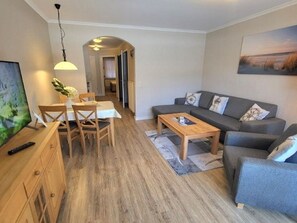 Uma sala moderna com um sofá cinzento, mesa de centro de madeira e área de refeições com mesa e cadeiras de madeira.
