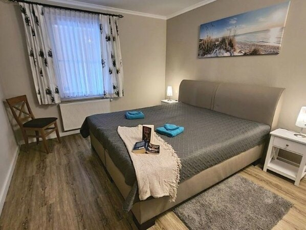 Quarto com cama, cadeira, mesa de cabeceira, abajur, janela com cortinas e um quadro na parede.