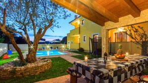 Un área junto a la piscina con un patio de piedra, una mesa dispuesta para comer y una vista de una casa amarilla al fondo.