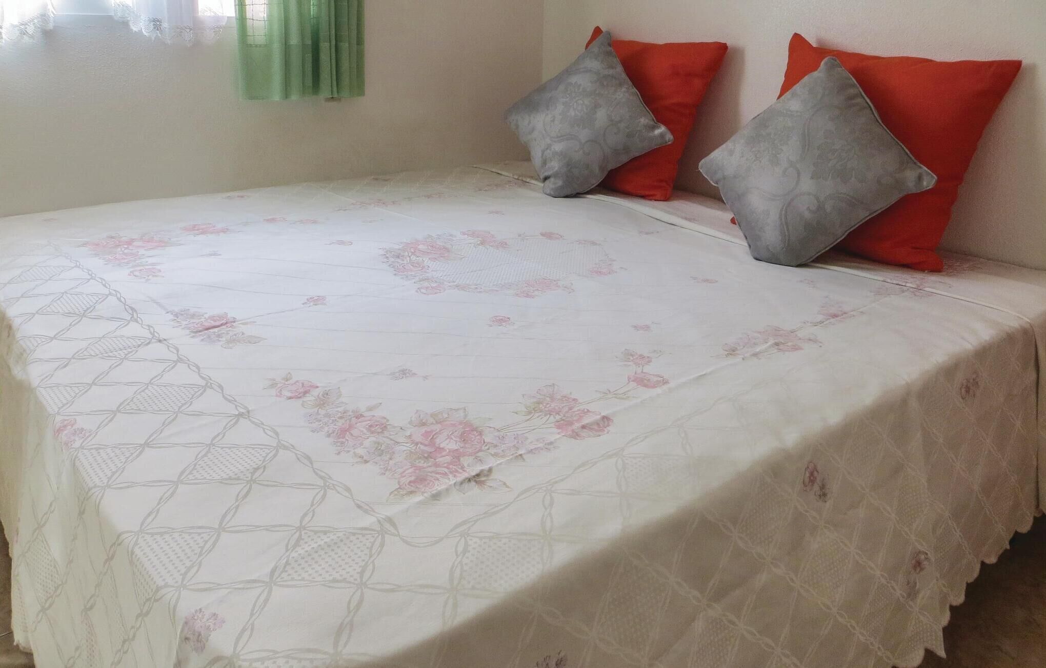 A bed with a floral-patterned bedspread and two pillows, one grey and one red, in a room with a plain wall and a window with green curtains.