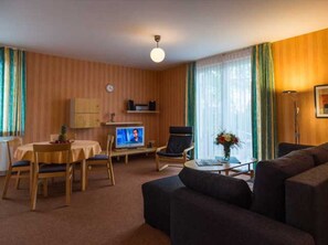 A living room with a sofa, a TV, a dining table with chairs, and a clock on the wall.
