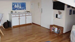 A compact living space with a kitchenette, a painting of sailboats, a television, and a small table with items on it. - (207) 2-room apartment - Appartementhaus Strandstr. 16 (Kühlungsborn)