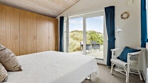 A bedroom with a bed, wooden wardrobe, white wicker chair, and a view of the outdoors. - Amazing home in Hvide Sande with sauna (Hvide Sande)