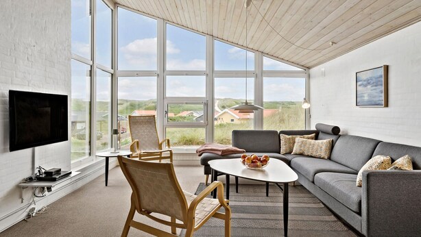 A spacious room with large windows, a grey sofa, a wooden chair, a small table with fruit, and a flat-screen TV mounted on the wall. - Amazing home in Hvide Sande with sauna (Hvide Sande)