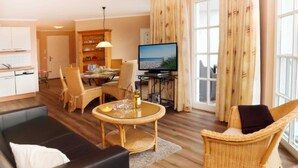 A living room with a sofa, a dining table with chairs, a television, and a kitchen area in the background. - (21) 3-room apartment - lake view - apartment house Atlantik (Kühlungsborn)
