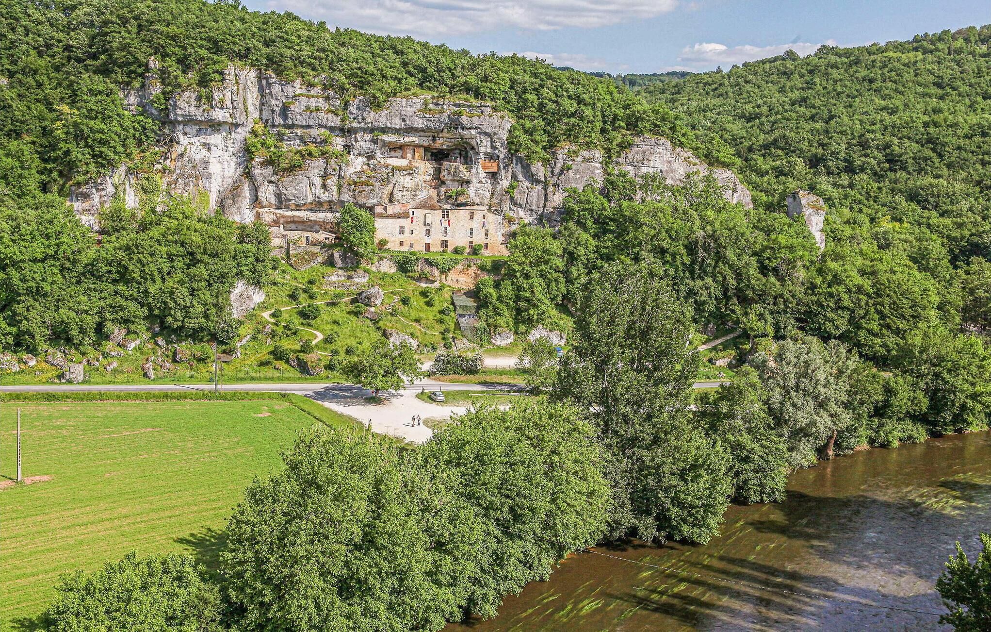 A historic stone building with a river in the foreground, surrounded by lush greenery and hills.