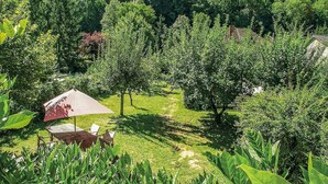 A lush garden with a wooden table and chairs under a red umbrella, surrounded by dense greenery and trees.