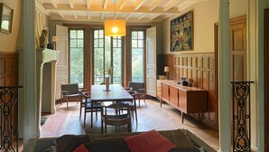 A dining area with a long table, chairs, a chandelier, and large windows.