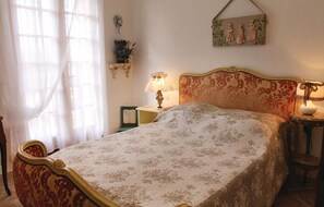 Een slaapkamer met een bed, nachtkastje, lamp, stoel, raam met gordijnen en een decoratie aan de muur.