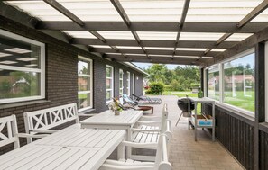 Ein überdachter Patio-Bereich mit Tischen, Stühlen, einem Grill und Blick auf einen Garten und Häuser.