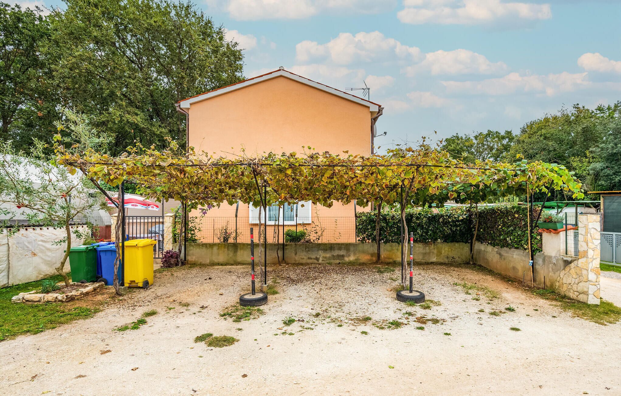 A building with a garden area featuring a grapevine pergola, a fenced yard, and a few bins.