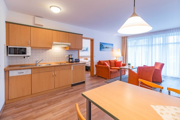 A modern kitchen with wooden cabinets, a microwave, and a dishwasher. There is a dining area with a table and chairs, and a sofa with orange cushions. A large window provides natural light.
